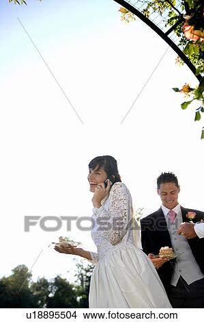 Picture - Young bride using cell phone, groom in background, smiling. Fotosearch