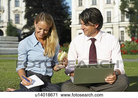 Stock Image - Businessman and businesswoman sitting in field working together.. Fotosearch