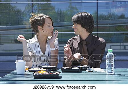 Stock Photo - Businessman and woman on table in front of office outside with sushi lunchbox and sticks she is eating he is watching her.. Fotosearch