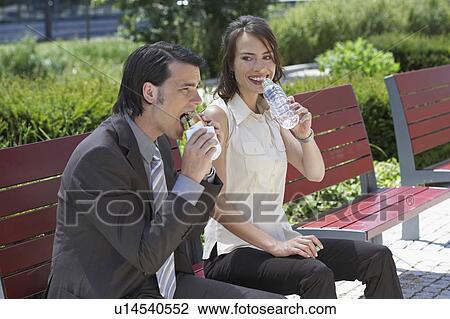 businessman and woman sitting on a bench outdoors she's about to drink water he is having a sandwich View Large Photo Image Stock Image - businessman and woman sitting on a bench outdoors she's about to drink water he is having a sandwich. Fotosearch