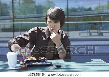 Picture - Businessman on table in front of office outside with sushi lunchbox and sticks licking off his finger.. Fotosearch