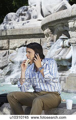 Stock Photography - Businessman sitting on edge of fountain eating sandwich whilst on the phone.. Fotosearch
