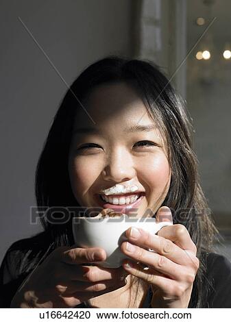Stock Image - Woman holding cappuccino, with foam on upper lip, smiling. Fotosearch