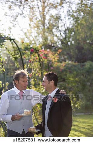 Stock Image - Mature father and groom holding plates of wedding cake in garden. Fotosearch