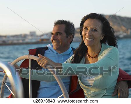 Stock Image - Mature couple at wheel of yacht, smiling. Fotosearch