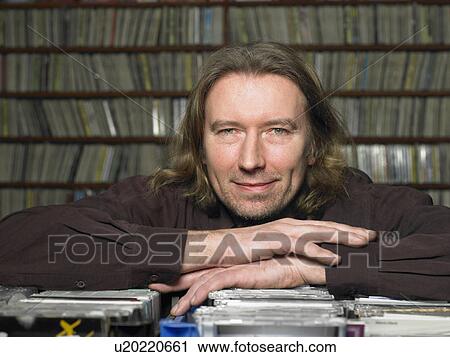 Stock Image - Mature man leaning on compact discs in shop, smiling, portrait. Fotosearch