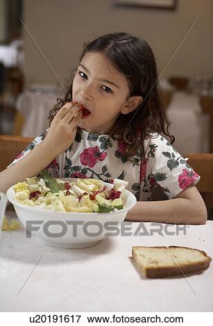 Stock Photo - Girl (5-7) eating salad from bowl at a table, portrait. Fotosearch
