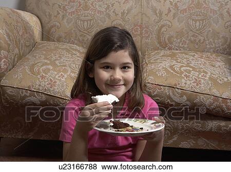 Stock Photo - Girl (6-8) sitting by sofa eating cake, smiling, portrait. Fotosearch