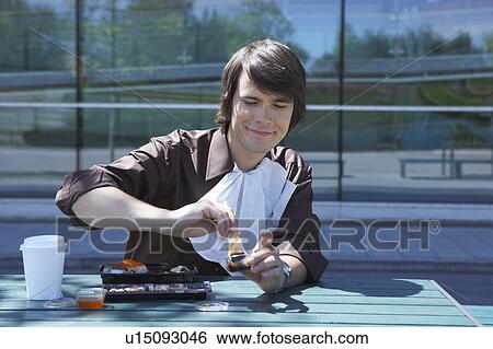 Stock Photograph - Smiling businessman eating sushi with chopsticks outside.. Fotosearch
