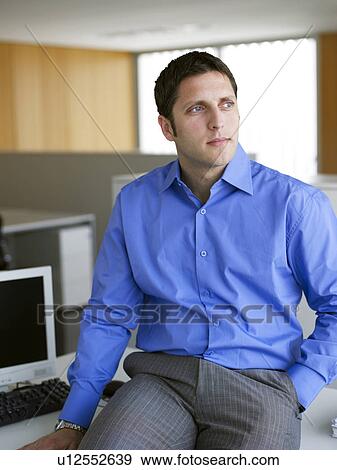 Young businessman sitting on desk in office View Large Photo Image Stock Photo - Young businessman sitting on desk in office. Fotosearch