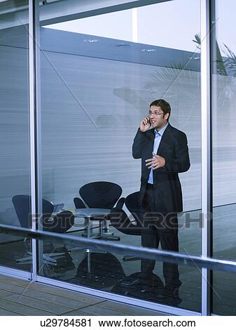 Young businessman standing at office window using mobile phone View Large Photo Image Stock Image - Young businessman standing at office window using mobile phone. Fotosearch