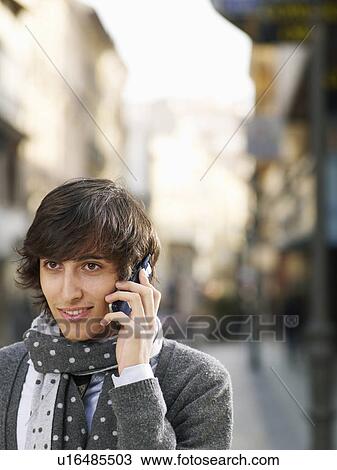 Stock Image - Young man standing in street using mobile phone, smiling, close-up. Fotosearch