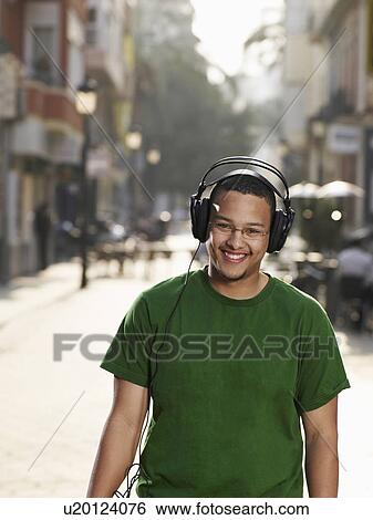 Young man standing in street wearing headphones, smiling, portrait View Large Photo Image Stock Photograph - Young man standing in street wearing headphones, smiling, portrait. Fotosearch