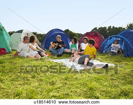 Stock Image - Group sitting outside tents one playing guitar. Fotosearch
