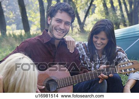 Man at campsite playing guitar with two woman listening and smiling. View Large Photo Image Picture - Man at campsite playing guitar with two woman listening and smiling.. Fotosearch