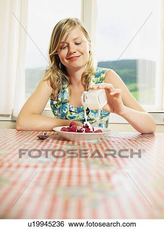 Stock Photograph - Woman pouring cream over bowl of raspberries at table smiling.. Fotosearch