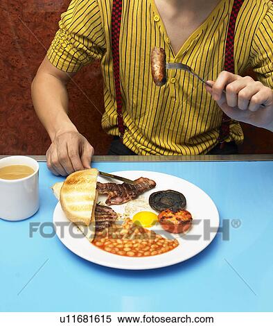 Stock Photography - Young man eating fried breakfast in cafe, mid section. Fotosearch