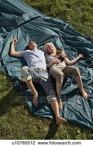 Stock Image - Couple lying on unraised tent smiling with the woman holding mallet.. Fotosearch