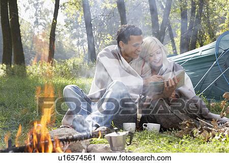 Stock Image - Couple sitting at campsite looking at map by the fire smiling.. Fotosearch