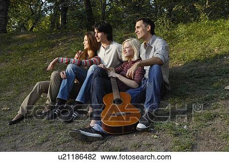 Stock Image - Four friends sitting with a guitar outdoors smiling.. Fotosearch