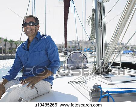 Stock Photograph - Mature man wearing sunglasses sitting on yacht, smiling. Fotosearch