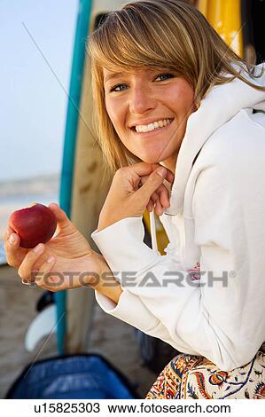 Stock Image - Woman eating an apple by surfboards smiling.. Fotosearch