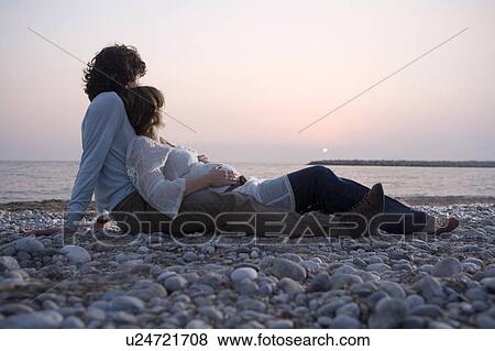 Stock Photo - Young couple lying on beach, woman pregnant, sunset. Fotosearch