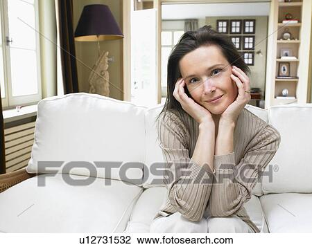 Stock Image - Woman sitting on sofa in living room.. Fotosearch