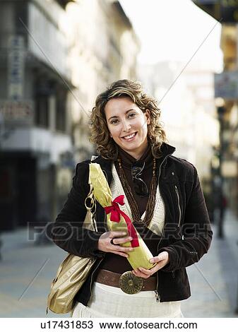 Stock Image - Young woman standing in street holding gift, smiling, portrait. Fotosearch