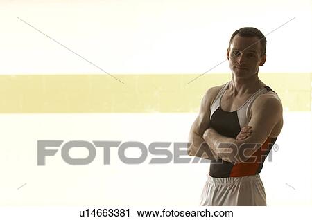 Stock Image - Male gymnast standing in gymnasium, arms crossed, portrait. Fotosearch