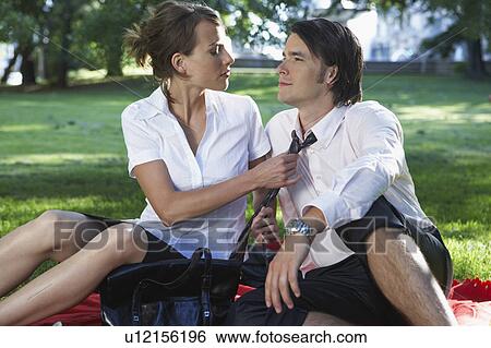 Stock Photograph - Businessman and woman sitting on blanket in meadow in park she is adjusting his tie.. Fotosearch