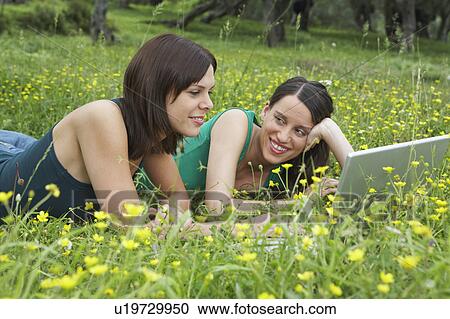 Stock Image - Two young women lying in meadow with laptop.. Fotosearch