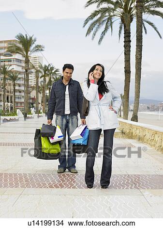 Woman using mobile phone, man carrying shopping in background View Large Photo Image Picture - Woman using mobile phone, man carrying shopping in background. Fotosearch