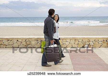Stock Photo - Young couple carrying shopping walking on promenade by beach. Fotosearch