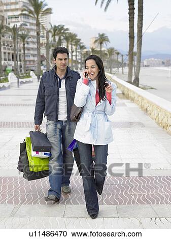 Stock Image - Couple walking along palm lined pavement, man carrying shopping bags while woman talks on mobile phone. Alicante, Spain.. Fotosearch