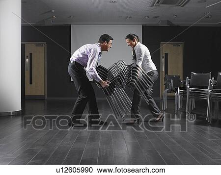 Stock Image - Man and woman moving stack of chairs in empty presentation room. Fotosearch