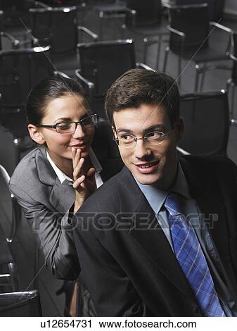 Stock Image - Woman whispering to man sitting in auditorium. Fotosearch