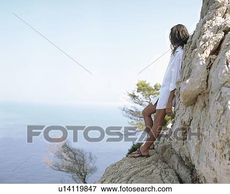 Stock Photo - Woman leaning on large rocks looking out over the sea.. Fotosearch