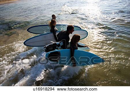 Picture - Four people carrying surfboards into the water laughing.. Fotosearch