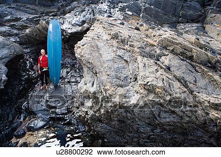 Man standing with surfboard on large rocks. View Large Photo Image Stock Image - Man standing with surfboard on large rocks.. Fotosearch