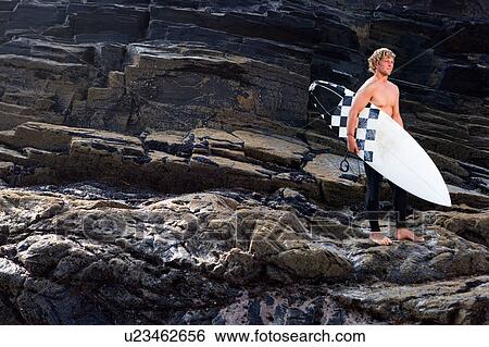Man standing with surfboard on large rocks. View Large Photo Image Stock Photograph - Man standing with surfboard on large rocks.. Fotosearch