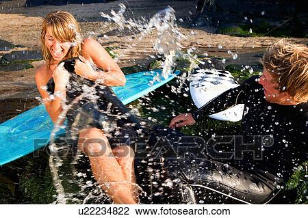 Stock Image - Couple splashing water on each other laughing with surfboards in background.. Fotosearch
