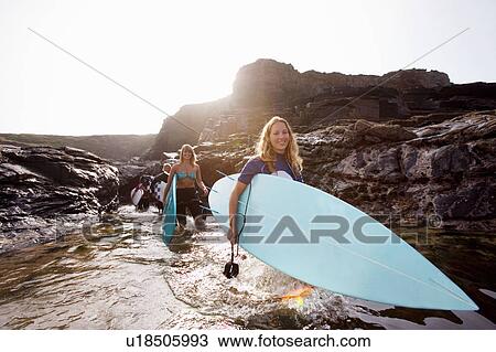 Stock Image - Four people carrying surfboards in the water smiling.. Fotosearch