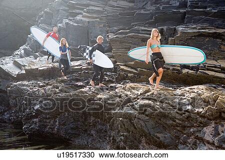 Stock Image - Four people carrying surfboards on large rocks.. Fotosearch