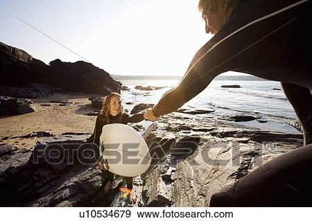 Stock Photo - Man helping woman with surfboard up large rocks smiling.. Fotosearch