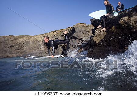 Man jumping into water on surfboard with two people sitting on large rocks smiling. View Large Photo Image Stock Photo - Man jumping into water on surfboard with two people sitting on large rocks smiling.. Fotosearch