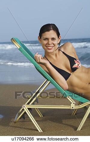 Stock Photograph - Young woman in bikini on beach in deck chair laughing into camera sea in background.. Fotosearch
