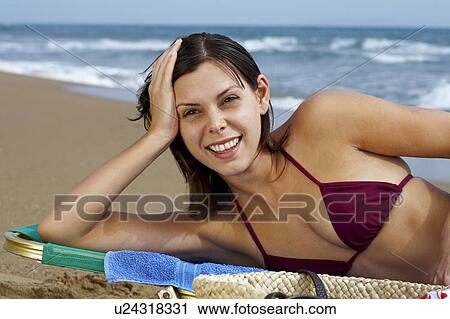 Stock Image - Young woman in bikini on beach lying in deck chair sea in background.. Fotosearch