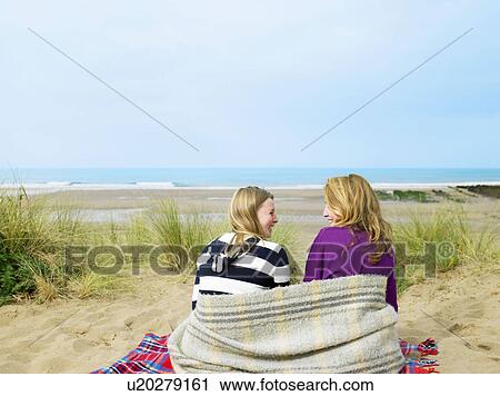 Stock Image - Girls sitting on sand dune with blanket. Fotosearch