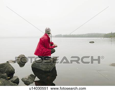Stock Image - Woman crouching on large rock in the water.. Fotosearch